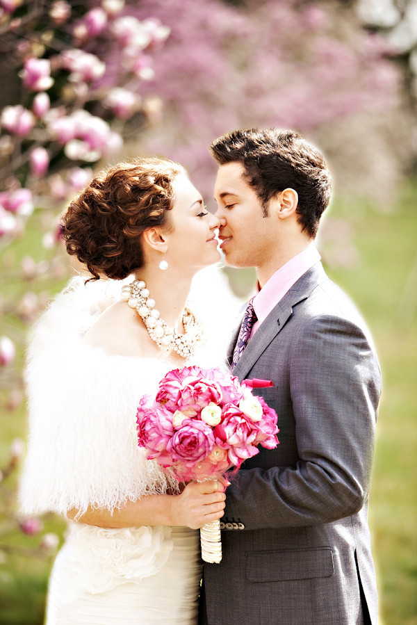 the bride and groom about to kiss - bride has hair in a loose updo and is wearing a white fur shawl, pearl and beaded necklace with matching earrings while holding a pink and white bouquet - groom is wearing a gray suit with a pink shirt - photo by North Carolina based wedding photographers Cunningham Photo Artists